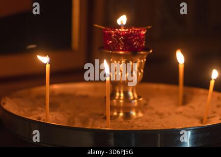 Eine ruhige Kirchenszene mit einem runden Sandtablett, mehreren beleuchteten Gebetskerzen und einem kunstvollen Goldhalter Stockfoto