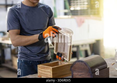 Schreiner hält und inspiziert eine Holzkiste in der Werkstatt. Zimmermann mit Sicherheitshandschuhen, die Holz in der Tischlerei bearbeiten Stockfoto