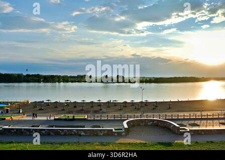 Flusspromenade bei Sonnenuntergang mit einem Sandstrand, Sonnenschirmen und Menschen, die entlang der gepflegten Terrassen entlang eines ruhigen, reflektierenden Flusses spazieren. Stockfoto