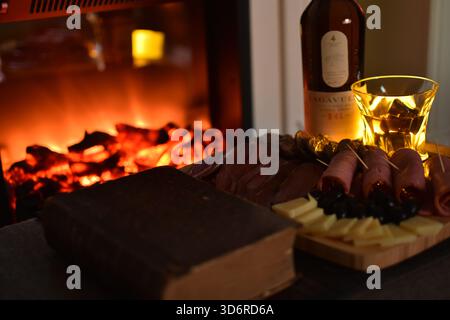 Lagavulin Single Malt Whisky Flasche und Glas am Kamin mit Fleisch- und Käsevorspeisen und einem Vintage Buch Stockfoto