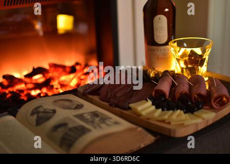 Lagavulin Single Malt Whisky Flasche und Glas am Kamin mit Fleisch- und Käsevorspeisen und einem Vintage Buch Stockfoto