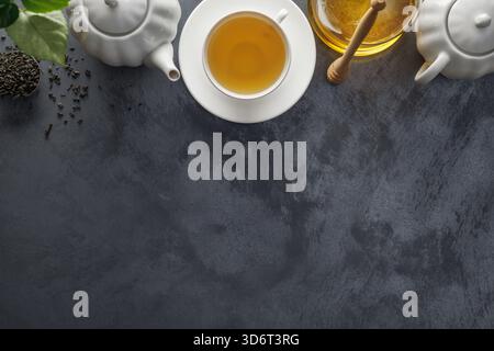Frischer Tee in einer weißen Keramikschale, Honig, Honiglöffel, Teekanne, Teeblätter auf dem Tisch. Stockfoto
