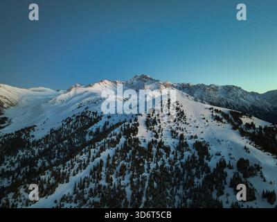 Blick aus der Vogelperspektive auf hohe, schneebedeckte Berggipfel und Bergketten mit teilweiser Sonneneinstrahlung, die den Schnee einfängt und über den dunklen Nadelwaldhang steigt Stockfoto