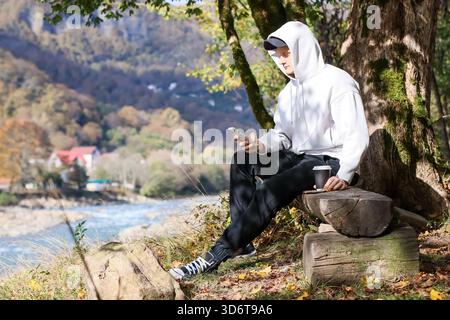 Mann in Kapuzenpullover durchsucht das Telefon mit Kaffee in der Hand, sitzt auf Holzholz am Fluss in der Herbstlandschaft. Stockfoto