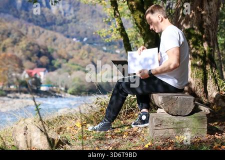 Ein Mann in legeren Kleidern untersucht weißes Papier, während er mit Laptop auf Holzholz in der Nähe des Herbstwaldes sitzt Stockfoto