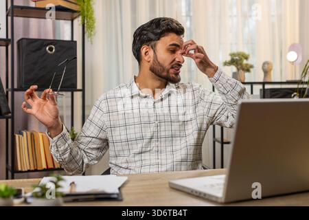 Überarbeiteter junger indischer bärtiger Geschäftsmann, der unter Augenanstrengung und Kopfschmerzen leidet und am Laptop arbeitet, der am Schreibtisch im Heimbüro sitzt. Müde Freelancer zieht Brillen und Massagen aus, reibt Nasenrücken. Stockfoto