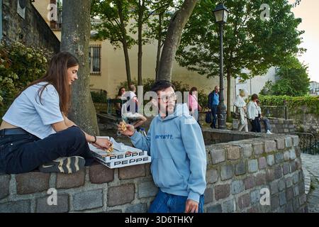 Paris, Frankreich - März 2025: Paare und Freunde treffen sich auf dem Platz Roland Dorgelès in Montmartre. Ein junger Mann und eine junge Frau teilen sich Pizza, während sie sich in der sonnen Stockfoto
