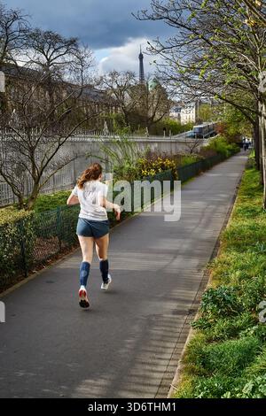 Paris, Frankreich - März 2025: Eine Frau joggt auf einem von Bäumen gesäumten Weg im 15. Pariser Arrondissement. Die Sonne scheint durch die Wolken und bildet eine Brig Stockfoto