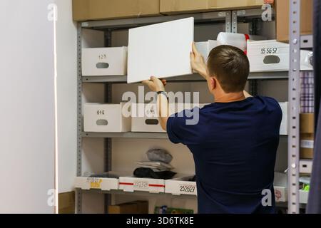 Rückansicht des Arbeiters in einem marineblauen T-Stück, das eine leere weiße Platte in das Lagerregal mit nummerierten Boxen einsetzt. Stockfoto