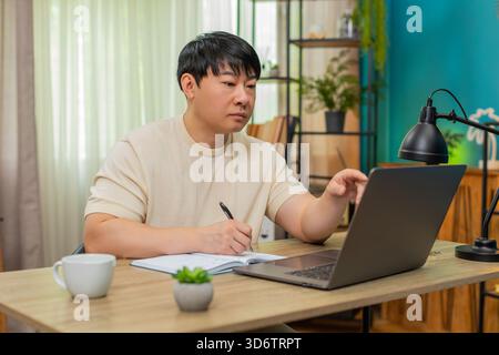Asiatischer Mann, der zu Hause am Tisch sitzt, nimmt an einem Online-Kurs auf Laptop Teil, schreibt Notizen und erklärt der Kamera, wie sie sich zeigt. Der Chinese spricht selbstbewusst, mit ausdrucksstarken Gesten, um ansprechende Online-Lektion zu erstellen. Stockfoto