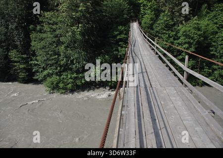 Eine lange Holzbrücke erstreckt sich über einen rauschenden Fluss, umgeben von dichten Bäumen und Laub. Sonnenlicht filtert durch das Grün und schafft ein ruhiges A Stockfoto