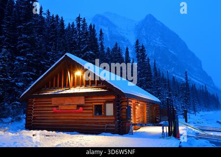 Eine Blockhütte steht am Rande eines gefrorenen Sees und unter den Berggipfeln, an einem verschneiten Winterabend in der Nähe der Weihnachtsfeiertage Stockfoto