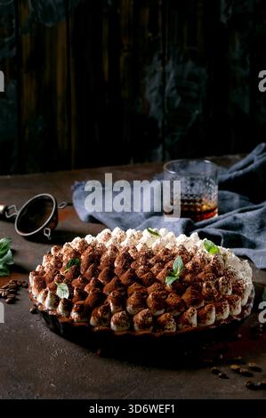 Hausgemachtes glutenfreies Tiramisu traditionelles italienisches Dessert mit Kakaopulver bestreut mit Minzblättern, Glas Whiskey, blauer Textilserviette und Kaffeebohnen auf dunklem Hintergrund. Stockfoto