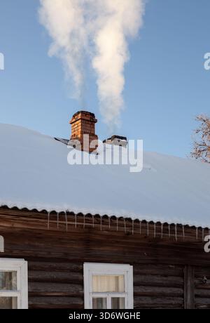Der Holzofen brennt in einem rustikalen Blockhaus. Eine Säule aus weißem Rauch erhebt sich aus einem gemauerten Kamin auf einem schneebedeckten Dach. Stockfoto