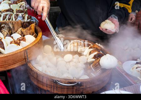 Nahaufnahme einer Hand, die eine Zange hält, die einen Teig aus einer Bambusschüssel mit Teigtaschen und gedämpften Brötchen an einem heißen Imbissstand in Chinatown entfernt Stockfoto