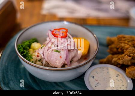 Nahaufnahme peruanischer Ceviche mit Fisch, Zwiebeln, Mais, Chili und Süßkartoffeln, serviert neben gebratenem Fisch und cremiger Sauce auf blauem Teller. Stockfoto
