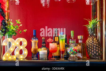 Farbenfrohe Cocktails, Spirituosenflaschen, Schüssel und dekorative Elemente auf einem festlichen Bartisch mit leuchtend roter Hintergrundbeleuchtung. Stockfoto