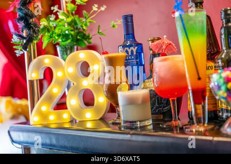 Farbenfrohe Cocktails, Spirituosenflaschen, Schüssel und dekorative Elemente auf einem festlichen Bartisch mit leuchtend roter Hintergrundbeleuchtung. Stockfoto