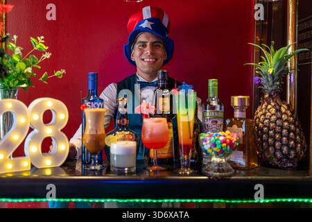 Festlicher Barkeeper hinter einer dekorierten Bar mit farbenfrohen Cocktails, Spirituosenflaschen, Schüssel und leuchtendem rotem Hintergrund für eine lebhafte Atmosphäre. Stockfoto