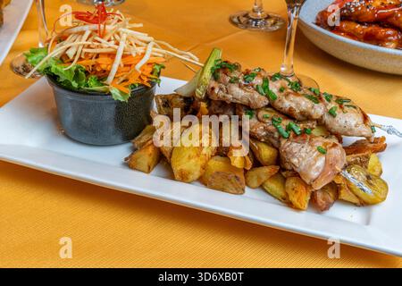 Gegrillte Hähnchenspieße, serviert mit gebratenen Kartoffeln und frischem Salat auf einem weißen Teller in einem Restaurant auf einer gelben Tischdecke mit Weinglas Stockfoto