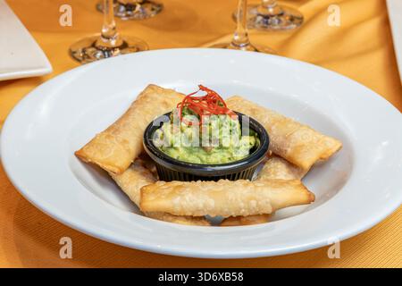 Tequenos-Käsestäbchen mit cremiger Guacamole auf einem weißen Teller über einem Restauranttisch mit heller, warmer Beleuchtung. Stockfoto
