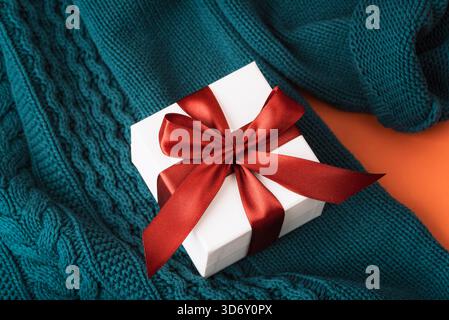 Eine weiße Geschenkbox mit rotem Band auf einem Strickpullover in blauem Blau mit weichen geflochtenen Nähten. Draufsicht. Stockfoto