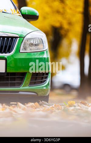 Grüne Fahrzeugfront im Herbst. Nahaufnahme eines hellgrünen Autos, das auf Straßenhöhe geparkt ist, mit einem weichen Vordergrund aus gefallenen gelben Blättern. Seaso Stockfoto