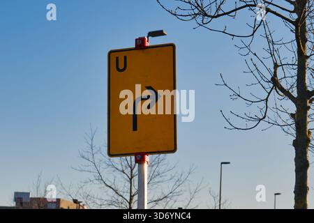 Ein gelbes Schild, das eine Rechtskurve anzeigt, ist vor einem blauen Himmel in Neuss deutlich sichtbar. Bäume und Straßenlaternen umrahmen die Szene und zeigen eine urb Stockfoto