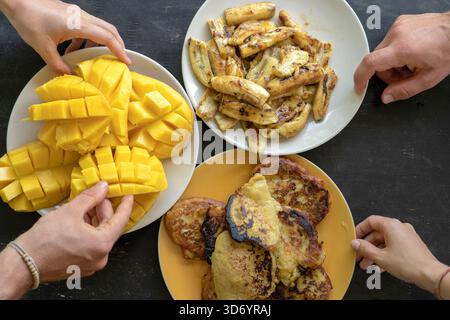 Bananenpfannkuchen, gebratene Bananen, gelbe Mango und Menschenhände. Leckeres Dessert, Nahaufnahme, Blick von oben. Gruppe glücklicher Freunde, die gutes Essen essen, genießen das Stockfoto