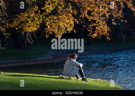Junger Mann, der am Duck Pond mit Skateboard im Boston Public Garden im Herbst sitzt Stockfoto