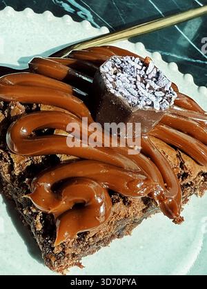 Nahaufnahme eines reichhaltigen Schokoladen-Brownie, gekrönt mit Schoko-Zuckerguss und einem dekorativen Schokoladen-Trüffel auf einem weißen Teller Stockfoto