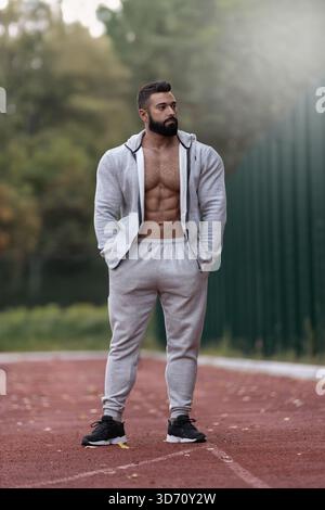 Muskulöser Mann steht auf einer Laufbahn in sportlicher Kleidung. Er scheint konzentriert und entspannt zu sein und hebt seine Fitness-Reise hervor. Der Hintergrund zeigt Bäume und einen klaren Himmel. Stockfoto
