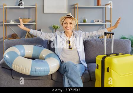 Glückliche blonde Frau, die entspannt auf einem bequemen Sofa mit gelbem Koffer und aufgeblasenem Ring sitzt Stockfoto