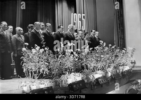 Slawjansk, Region Donezk, ukrainische SSR, Sowjetunion. 1965. Die Mitglieder des Präsidiums stehen auf der Bühne des Lenin-Kulturpalastes während der Feier zum 50. Jahrestag der AIZ-Fabrik. Eine Gruppe ernster Männer in dunklen Anzügen und ein Militäroffizier stehen hinter einer langen Reihe von Topfblumen auf der Hut. Das Datum 1965 hängt auf dem Vorhang nahe einer Leninbüste. Dieses Schwarzweißfoto im Archiv zeigt die offizielle Atmosphäre der sowjetischen Feierlichkeiten in der friedlichen Donbass-Region lange vor dem Krieg. Stockfoto
