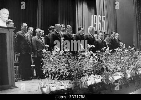 Slawjansk, Region Donezk, ukrainische SSR, Sowjetunion. 1965. Die Mitglieder des Präsidiums stehen auf der Bühne des Lenin-Kulturpalastes während der Feier zum 50. Jahrestag der AIZ-Fabrik. Eine Gruppe ernster Männer in dunklen Anzügen und ein Militäroffizier stehen hinter einer langen Reihe von Topfblumen auf der Hut. Das Datum 1965 hängt auf dem Vorhang nahe einer Leninbüste. Dieses Schwarzweißfoto im Archiv zeigt die offizielle Atmosphäre der sowjetischen Feierlichkeiten in der friedlichen Donbass-Region lange vor dem Krieg. Stockfoto