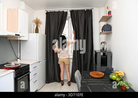 Eine 50-jährige Russin öffnet dunkle Vorhänge in einer gemütlichen Küche, beginnt einen neuen Tag und bereitet sich in ruhiger Morgenatmosphäre auf das Frühstück vor. Stockfoto