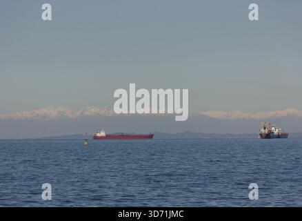 Frachtschiff, das auf ruhigem Meer mit weit entfernten schneebedeckten Bergen am Horizont segelt Stockfoto