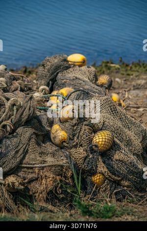 Nahaufnahme alter, verworrener brauner Fischernetze mit hellgelben Schwimmern, die sich am grasbewachsenen Ufer in der Nähe von ruhigem, blauem Wasser stapelten Stockfoto