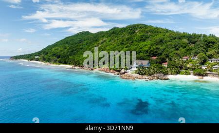 Panoramablick auf üppige grüne Hügel, die unter klarem Himmel in türkisfarbenes Wasser der Küste absteigen. Anse Patates. Seychellen, La Digue. Stockfoto