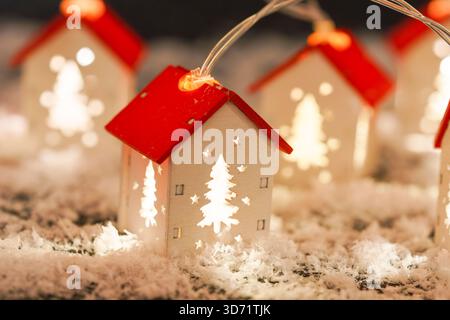 Eine Schnur aus leuchtenden Miniaturlichtern. Weiße Weihnachtshäuser mit rotem Dach auf künstlichem Schnee Hintergrund, warme und gemütliche Winterferienszene Stockfoto