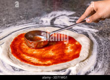 Nahaufnahme Hand von Koch Bäcker, der Pizza in der Küche macht. Der Koch gießt Tomatensauce über den gerollten Teig. Chef Hand verteilt pasteurisierte Tomatenpaste Stockfoto