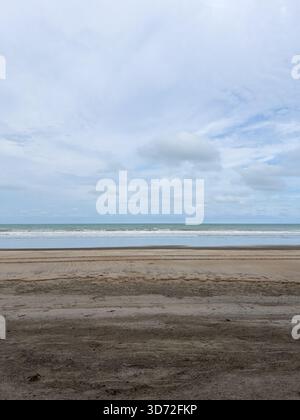 Ein Foto im vertikalen Format, das eine weite Weite aus feuchtem, dunklem Sand im Vordergrund zeigt und auf die sanften Wellen des blau-grünen Ozeans trifft Stockfoto