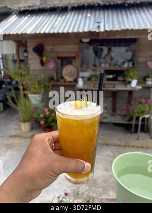 Kuantan, Pahang, Malaysia - 23. November 2025 : Eine Hand, die ein hohes Glas eines orange-gelben Eisgetränks hält (wahrscheinlich Eistee, Eiskaffee oder Mocktail) Stockfoto