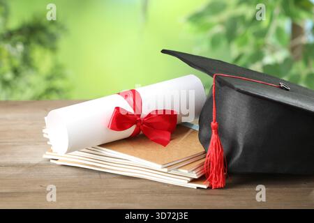 Graduierungskappe, Diplom und Notizbücher auf Holztisch vor verschwommenem Hintergrund, Großaufnahme Stockfoto