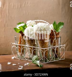 Hausgemachte süße Waffelkegel mit weißen Blumen und grünen Blättern, in transparenten Gläsern auf Holztisch. Sommer natürliche Blumensüße Desserts Konzept. Kopierbereich Stockfoto