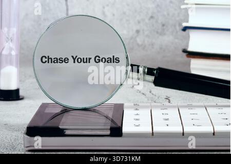 Eine Lupe hebt den Satz „Chase Your Goals“ auf einem Rechner hervor, mit einer Sanduhr und einem Stapel Bücher im Hintergrund. Konzentrieren Sie sich auf Stockfoto