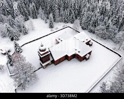 Aus der Vogelperspektive der hölzernen Gelenkkirche von Svaty Kriz in der Region Liptov, die neben einem dichten verschneiten Wald steht und von der ruhigen rur umgeben ist Stockfoto