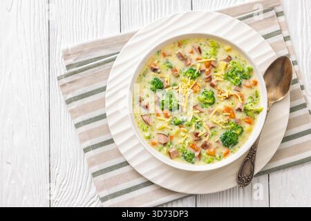Wurstgemüse Chowder, dicke cremige Suppe mit Brokkoli, Karotten, Kartoffeln in weißer Schüssel auf weißem strukturiertem Holztisch mit Serviette und Löffel, Stockfoto