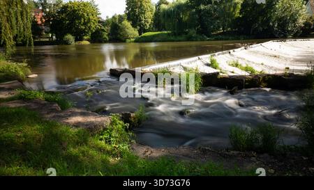 Ein breiter Blick auf einen Fluss oder Bach, der über ein kleines Steinwehr fließt, umgeben von üppigen, üppigen grünen Bäumen und Laub, einschließlich eines Weinwillens Stockfoto