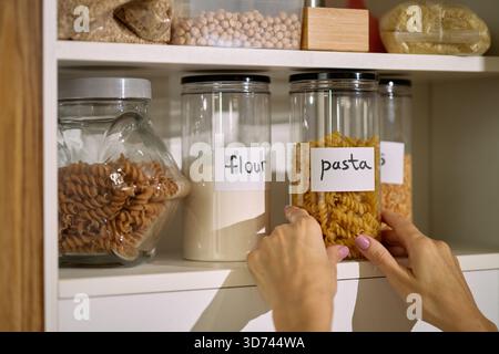 Kaukasische Frau, die die Küchenküche organisiert, ein Glas Pasta auf ein Regal neben Mehl- und andere Trockenwaren legt, nur Hände und Teile der Arme sind aus der Nähe sichtbar Stockfoto